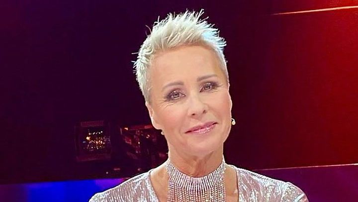 Sonja Zietlow mit kurzen, blondierten Haaren und einem eleganten silbernen Outfit sitzt in einem Studio mit bunten Lichteffekten im Hintergrund. Sie trägt ein auffälliges Halsband und hat ein freundliches Lächeln im Gesicht. Sonja Zietlow mit kurzen, blondierten Haaren und einem eleganten silbernen Outfit sitzt in einem Studio mit bunten Lichteffekten im Hintergrund. Sie trägt ein auffälliges Halsband und hat ein freundliches Lächeln im Gesicht.