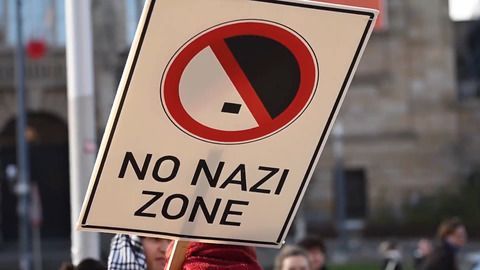 Ein Demonstrationsschild mit der Aufschrift „NO NAZI ZONE“ in schwarzen Großbuchstaben. Darüber befindet sich ein rot durchgestrichenes Symbol, das an eine stilisierte Fahne erinnert. Im Hintergrund sind unscharfe Gebäude und Personen zu erkennen. Ein Demonstrationsschild mit der Aufschrift „NO NAZI ZONE“ in schwarzen Großbuchstaben. Darüber befindet sich ein rot durchgestrichenes Symbol, das an eine stilisierte Fahne erinnert. Im Hintergrund sind unscharfe Gebäude und Personen zu erkennen.